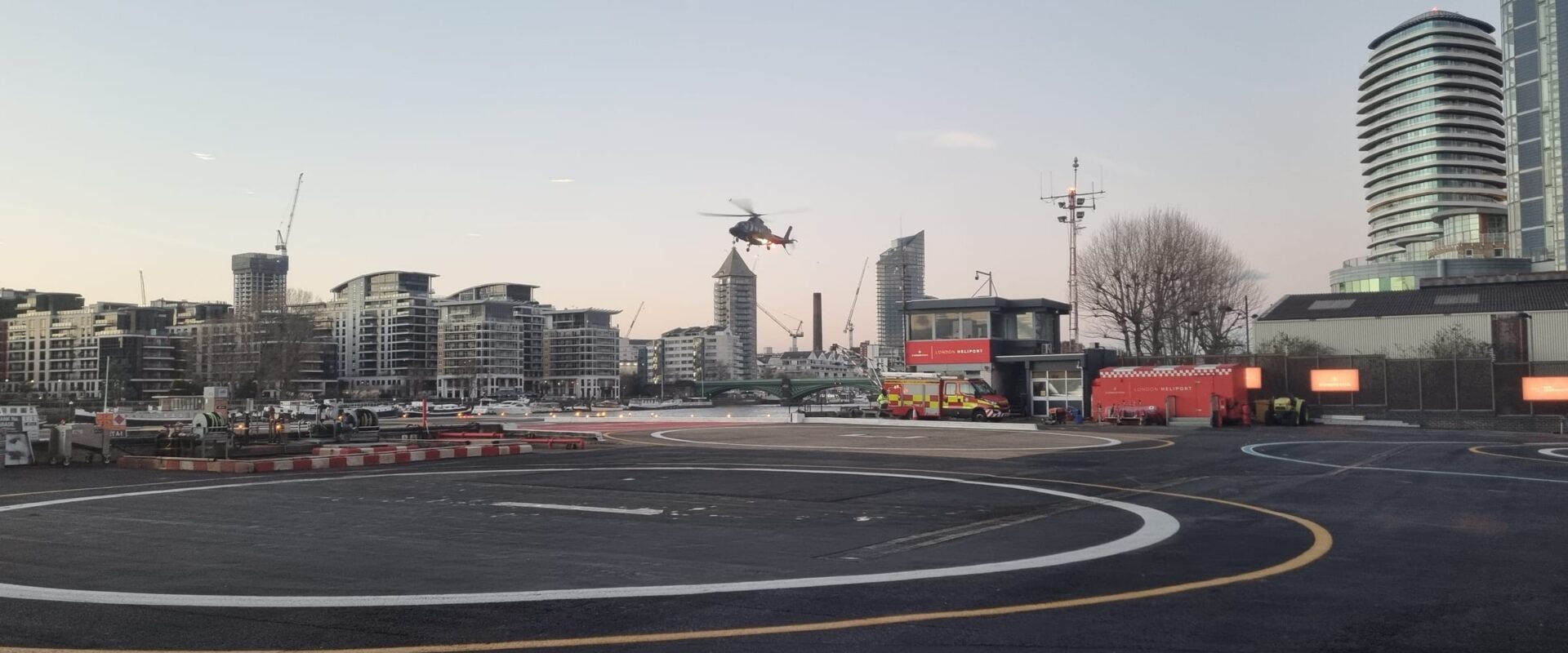 A helicopter arriving over the landing pad at London Heliport in Battersea at dusk, with the control tower and the Thames riverside behind