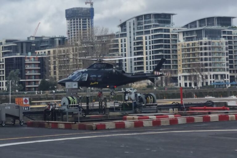 A black twin-engine Agusta 109 helicopter beside the fuelling area at London Heliport in Battersea, with riverside flats behind