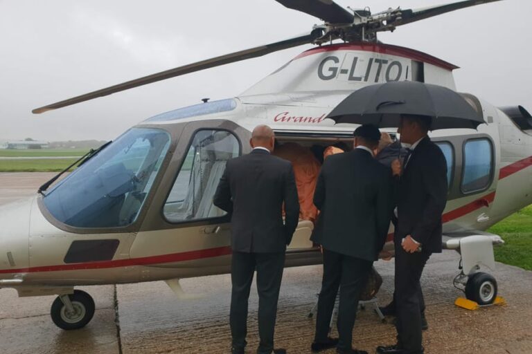 Three passengers in dark suits boarding an Agusta Grand helicopter on a wet day, one sheltering under an umbrella